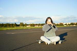 branché adolescent fille séance sur sa planche à roulette, prise Photos sur téléphone intelligent. asiatique femme patineur est assis sur longboard et photographier sur mobile téléphone