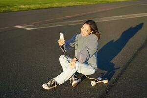 magnifique coréen fille prend selfie sur téléphone intelligent, prend photo avec sa planche à roulette, apprécie ensoleillé journée en plein air