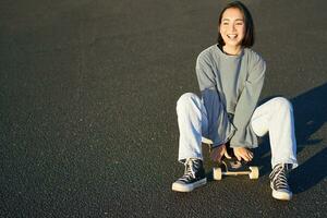 liberté et Bonheur. mignonne souriant asiatique fille, est assis sur planche à roulette sur ensoleillé printemps journée. content en riant patineur profiter croisière sur longboard photo