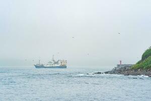 pêche navire émerge de derrière une cap avec une phare, voile dans une brumeux mer photo