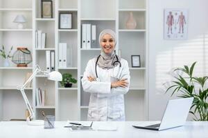 portrait de Jeune femelle médecin portant hijab, musulman femme souriant et à la recherche à caméra avec bras franchi dans blanc médical manteau avec stéthoscope, travail à l'intérieur médical Bureau bureau.. photo