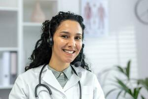 portrait de Jeune magnifique femelle médecin avec casque téléphone vidéo appel, hispanique femme souriant et à la recherche à caméra proche en haut, ouvrier travail à l'intérieur en ligne clinique Bureau consultant les patients. photo