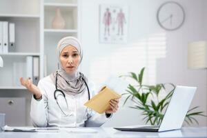 portrait de une triste musulman arabe femelle médecin séance à une table dans un Bureau dans une privé clinique, en portant un enveloppe, mal nouvelles, décevant tester résultats, à la recherche à le caméra. photo