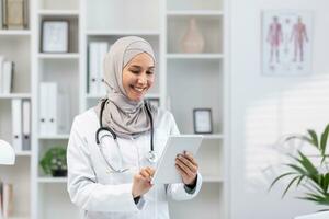 Jeune magnifique femelle médecin dans hijab en utilisant tablette ordinateur, médecin ouvrier dans blanc médical manteau et stéthoscope à l'intérieur clinique Bureau à lieu de travail, souriant musulman femme en utilisant en ligne application. photo