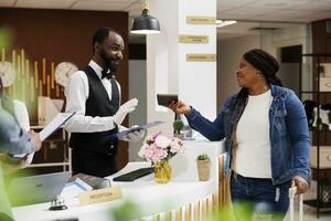 Jeune africain américain femme touristique montrant passeport à Hôtel Personnel pendant enregistrement processus à accueil comptoir, souriant femelle voyageur avec bagage enregistrement à de face bureau. client enregistrement photo