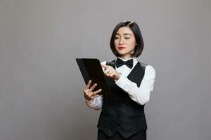 asiatique femme réceptionniste tapoter sur numérique tablette écran tactile tandis que travail dans hospitalité industrie. restauration un service serveuse dans noir et blanc uniforme vérification commande en ligne photo