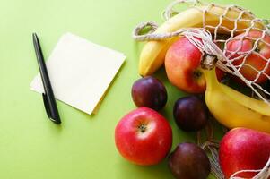 rouge pommes, bananes, prunes, chaîne sac et Vide papier avec noir stylo sur une brillant vert Contexte. photo