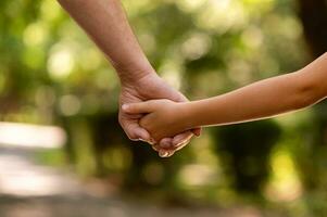 le du père main tenue fermement le main de le sien peu fils contre le Contexte de vert feuillage, fermer photo