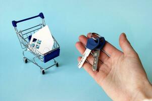 une main détient le clés et suivant à il est une mini-chariot avec une maison fabriqué de blanc papier. photo