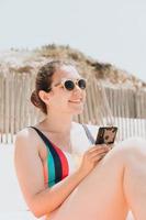 jeune femme sourit en discutant au téléphone portant un maillot de bain coloré à la plage en prenant un bain de soleil, concept de jeune vacances de voyage, espace de copie, réseau social, concept d'utilisateur de lunettes de soleil, gros plan
