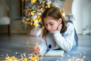 une mignonne fille mensonges sur le sol près le Noël arbre et écrit une lettre à Père Noël claus photo