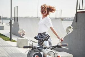 portrait d'une belle fille hipster assise sur un scooter rétro noir, souriant posant et profitant du chaud soleil du printemps. photo