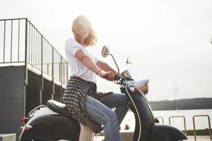 portrait d'une belle fille hipster assise sur un scooter rétro noir, souriant posant et profitant du chaud soleil du printemps. photo