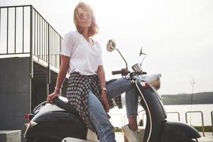 portrait d'une belle fille hipster assise sur un scooter rétro noir, souriant posant et profitant du chaud soleil du printemps. photo