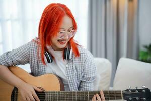 asiatique femme portant des lunettes et casque de musique en jouant guitare tandis que séance sur canapé dans le vivant pièce à maison. asiatique femmes l'écriture chanson tandis que en jouant guitare à maison. composer chanson la musique concept. photo
