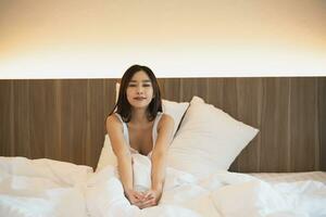 asiatique femme pose sur le lit, élongation et content femme veille en haut dans le Matin après dormir, du repos et se détendre à maison. repos, sourire et étendue dans chambre à coucher, pacifique, de bonne humeur confortable concept. photo