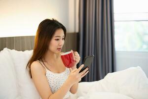 joyeux asiatique femme boisson chaud café et en utilisant mobile téléphone tandis que séance sur le lit dans chambre à maison. asiatique femmes relex pose sourire après réveiller en haut sur le lit maison. activité loisir à maison concept. photo