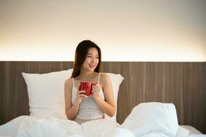 joyeux asiatique femme boisson chaud café tandis que séance sur le lit dans chambre à maison. asiatique femmes relex pose sourire après réveiller en haut sur le lit maison. activité loisir à maison concept. photo