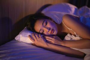 asiatique femme en train de dormir dans confortable lit avec soyeux linge de maison à nuit lumière. femmes mensonge dans lit et en gardant yeux fermé tandis que couvert avec couverture. contenu et pacifique, de bonne humeur et confortable. photo