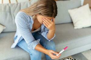 triste, inquiet femme vérification sa récent grossesse test, séance sur canapé à maison. maternité, enfant naissance et famille problèmes concept. indésirable grossesse photo