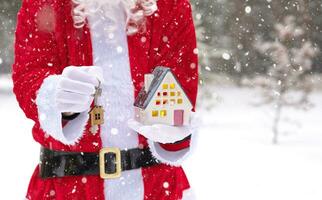 Père Noël soigneusement détient dans le sien mains minuscule maison dans petit Taille et clé avec une porte-clés dans neige Extérieur - une cadeau pour Noël, Nouveau année, rêve. protection, assurance, hypothèque, achat de logement photo