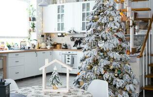 de fête Noël décor dans blanc cuisine, moderne village intérieur avec une neigeux Noël arbre et Fée lumières. Nouveau année, Noël humeur, confortable maison, métal modulaire escalier. le général plan photo