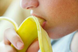 portrait de une adolescent garçon en mangeant une banane. photo de une gars bouche et lèvres avec une banane. Frais fruit. en bonne santé nourriture pour les enfants