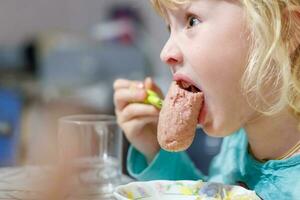 une peu fille a petit déjeuner à Accueil spaghetti avec saucisses. peu blond fille en mangeant dîner avec fourchette à table photo