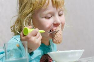 une peu fille a petit déjeuner à Accueil spaghetti avec saucisses. peu blond fille en mangeant dîner avec fourchette à table photo