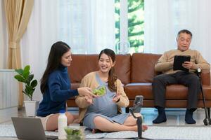 une Enceinte asiatique femme est assis sur le sol avec sa plus jeune sœur. vivre social médias diffuser vente Enceinte des mères des produits et repas, là est une père dans le Contexte relaxant sur une canapé. photo