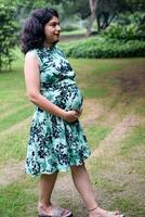 une Enceinte Indien Dame pose pour Extérieur grossesse tirer et mains sur ventre, Indien Enceinte femme met sa main sur sa estomac avec une maternité robe à société parc, Enceinte à l'extérieur maternité tirer photo