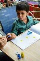 intelligent Indien peu garçon effectuer pouce La peinture avec différent coloré l'eau Couleur trousse pendant le été les vacances, mignonne Indien enfant Faire coloré pouce La peinture dessin sur en bois table photo