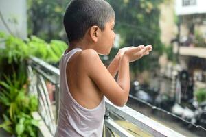 peu enfant en jouant dans été pluie dans maison balcon, Indien intelligent garçon en jouant avec pluie gouttes pendant mousson pluvieux saison, enfant en jouant dans pluie photo