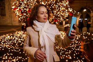 magnifique femme à fête foraine, en portant mobile téléphone, prise une soi portrait contre illuminé Noël arbre dans le nuit photo