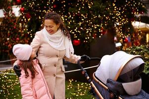 content souriant Jeune femme, aimant mère à la recherche à sa fille, pousser bébé landau, prendre plaisir Noël atmosphère en plein air photo