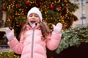 souriant content fille dans hiver vêtements, sourit à caméra, permanent suivant à illuminé Noël arbre à Noël juste photo