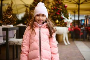 charmant content peu enfant fille sourit à caméra, permanent en plein air à Noël équitable, profiter le A venir de fête événements photo