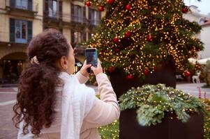 arrière vue femme photographier le illuminé Noël arbre sur sa téléphone intelligent tandis que en marchant le long de italien ville des rues photo