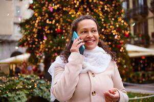 content femme parlant sur mobile téléphone, souriant contre illuminé Noël arbre Contexte à famille traditionnel marché photo