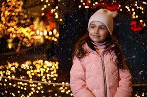 adorable enfant fille sourit rêveusement à la recherche loin, permanent Extérieur contre allégé rue et Noël arbre à fête foraine photo