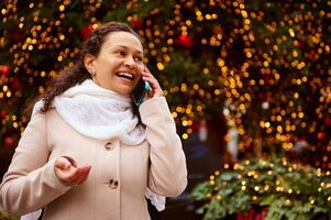 souriant femme parlant sur mobile téléphone contre illuminé Noël arbre Contexte photo