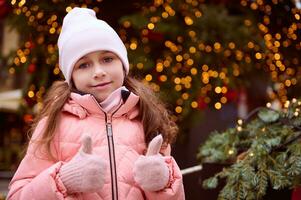 mignonne peu fille ayant amusement à Noël équitable, montrant les pouces en haut à caméra, permanent par illuminé Noël arbre photo