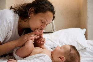 fermer Jeune aimant soins maman doucement baisers peu pieds et minuscule les orteils de sa nouveau née bébé garçon, profiter dépenses soumissionner des moments avec sa bien-aimée enfant. mère et enfant connexion. bébé se soucier concept photo