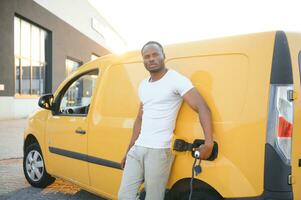 une africain américain homme des stands suivant à Jaune électrique livraison van à électrique véhicule mise en charge stations photo