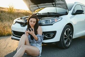 le Jeune fille cassé le voiture et elle ouvert le capuche et a essayé à réparation le voiture sur le route. une femme appels sur une téléphone intelligent avec une voiture évacuation un service photo