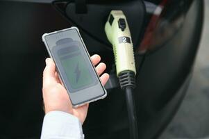 une homme des charges un électrique voiture et chèques le charge niveau sur une téléphone intelligent photo