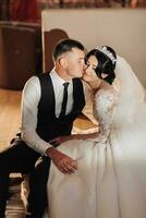 mariage portrait de le la mariée et jeune marié. intérieur la photographie avec magnifique lumière. élégant jeune marié dans une noir gilet baisers une brunette la mariée sur le joue, séance étreindre à l'intérieur. couple dans l'amour photo