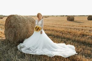 mariage portrait. une roux la mariée dans une blanc robe, en portant une bouquet de tournesols, sur le Contexte de une champ. magnifique boucles. sincère sourire. élégant robe. Jaune fleurs photo