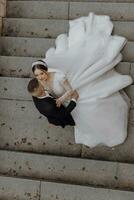 charmant la mariée dans une magnifique robe, une longue voile, le jeune marié virages le la mariée comme si dans une danse. une mariage couple dans le ville centre sur le escaliers. photo de au dessus