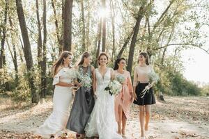 mariage photo dans la nature. le la mariée et demoiselles d'honneur sont en marchant dans le forêt souriant, en portant leur bouquet et à la recherche à le la mariée. content mariage concept. émotions. les filles